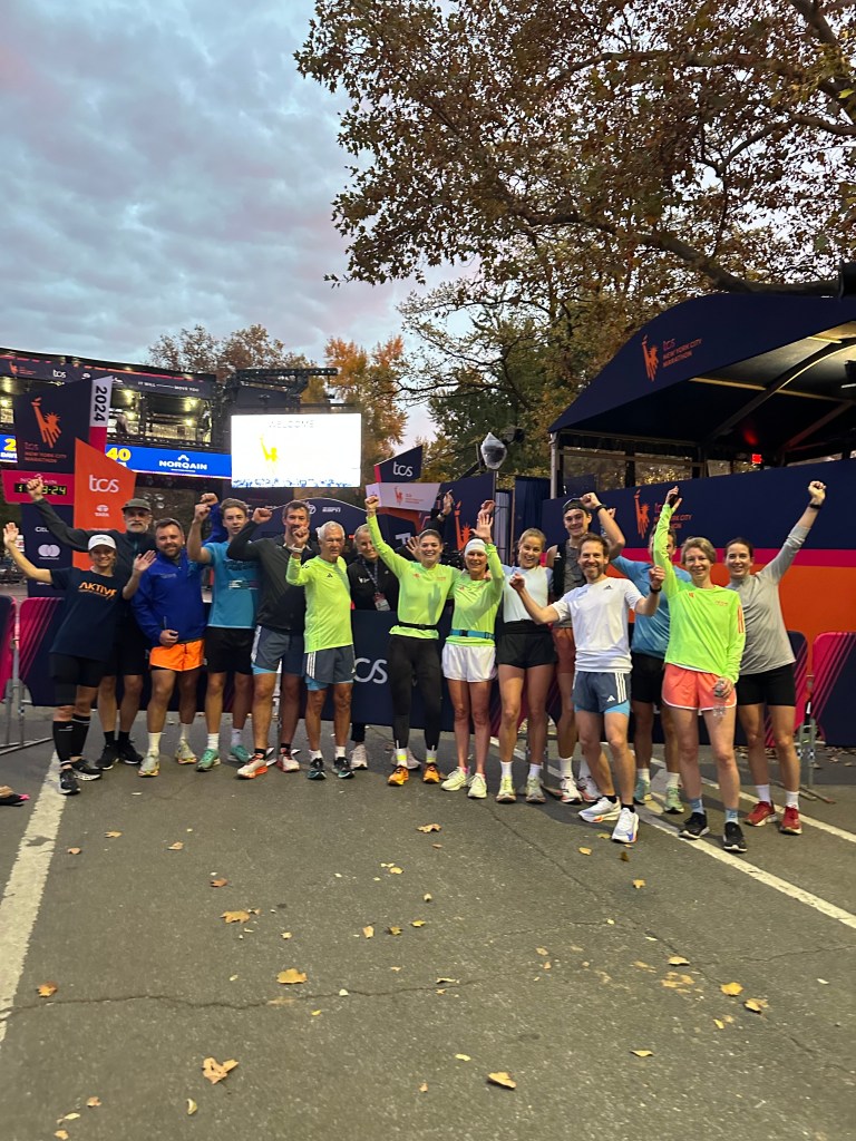 Gruppe med glade deltakere som feirer etter TCS New York City Marathon, ikledd fargerike treningsklær og med hevede hender på et område med høstlige blader.