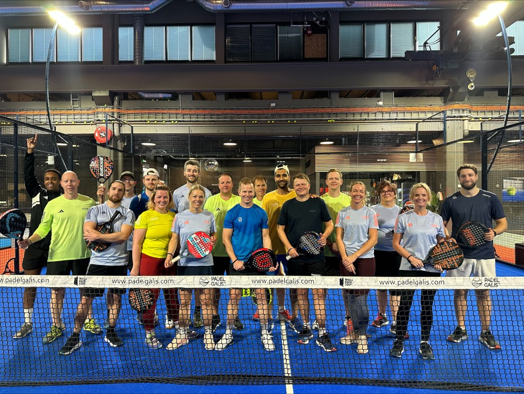 Gruppe av mennesker som poserer sammen på en padelbane, iført sportsklær og holder padelracketer.