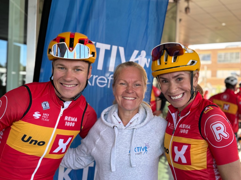 Tre personer smiler på et arrangement, iført sykkeldrakter og hjelmer, med en 'Aktiv mot kreft'-banner i bakgrunnen.