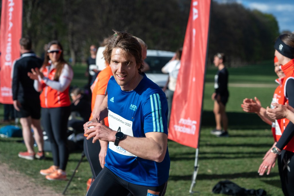 En deltaker smiler mens han løper ved et arrangement, omringet av tilskuere som heier, med bannere i bakgrunnen.