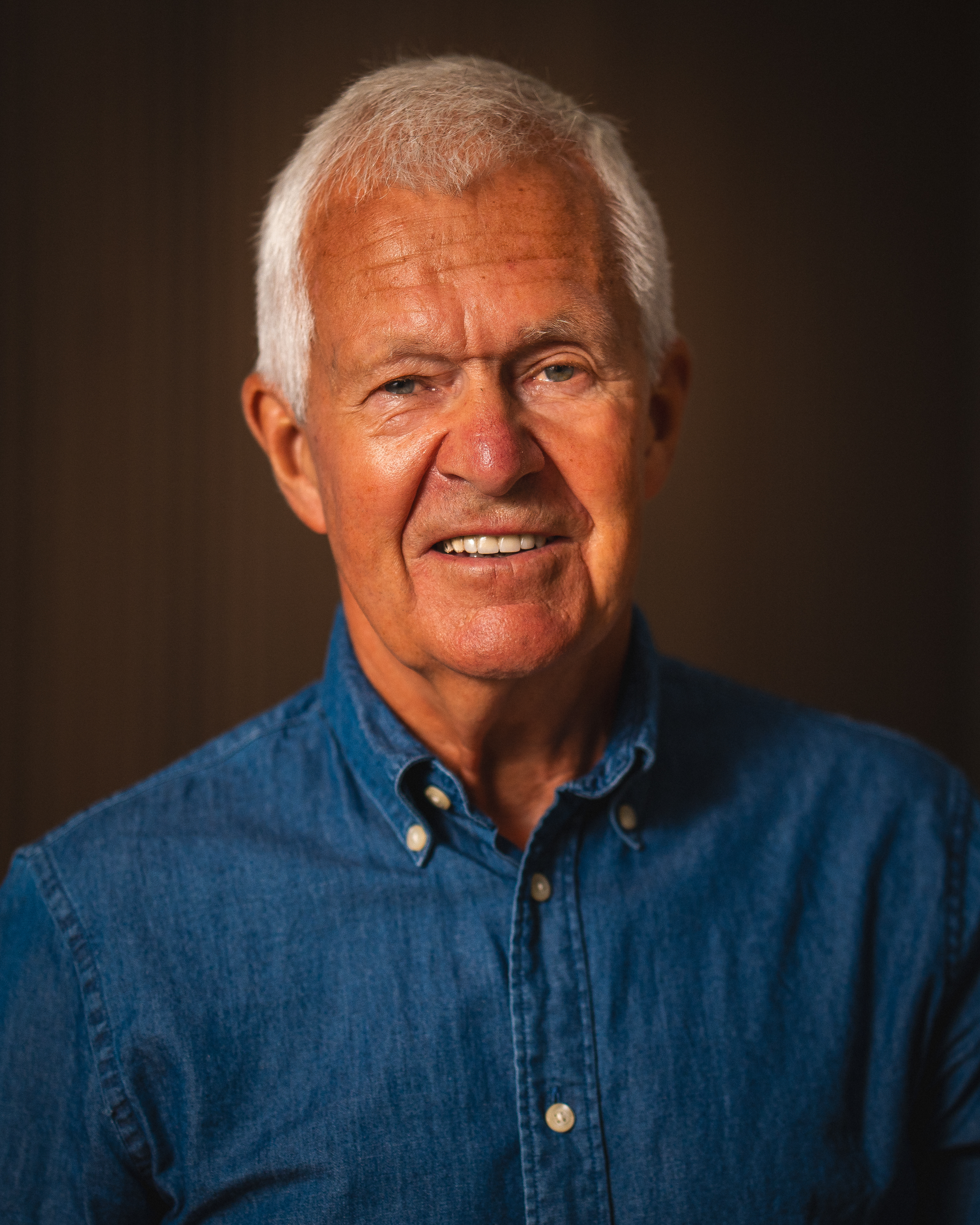 Portrett av en eldre mann med kort, grått hår og smilende uttrykk, iført en blå skjorte.