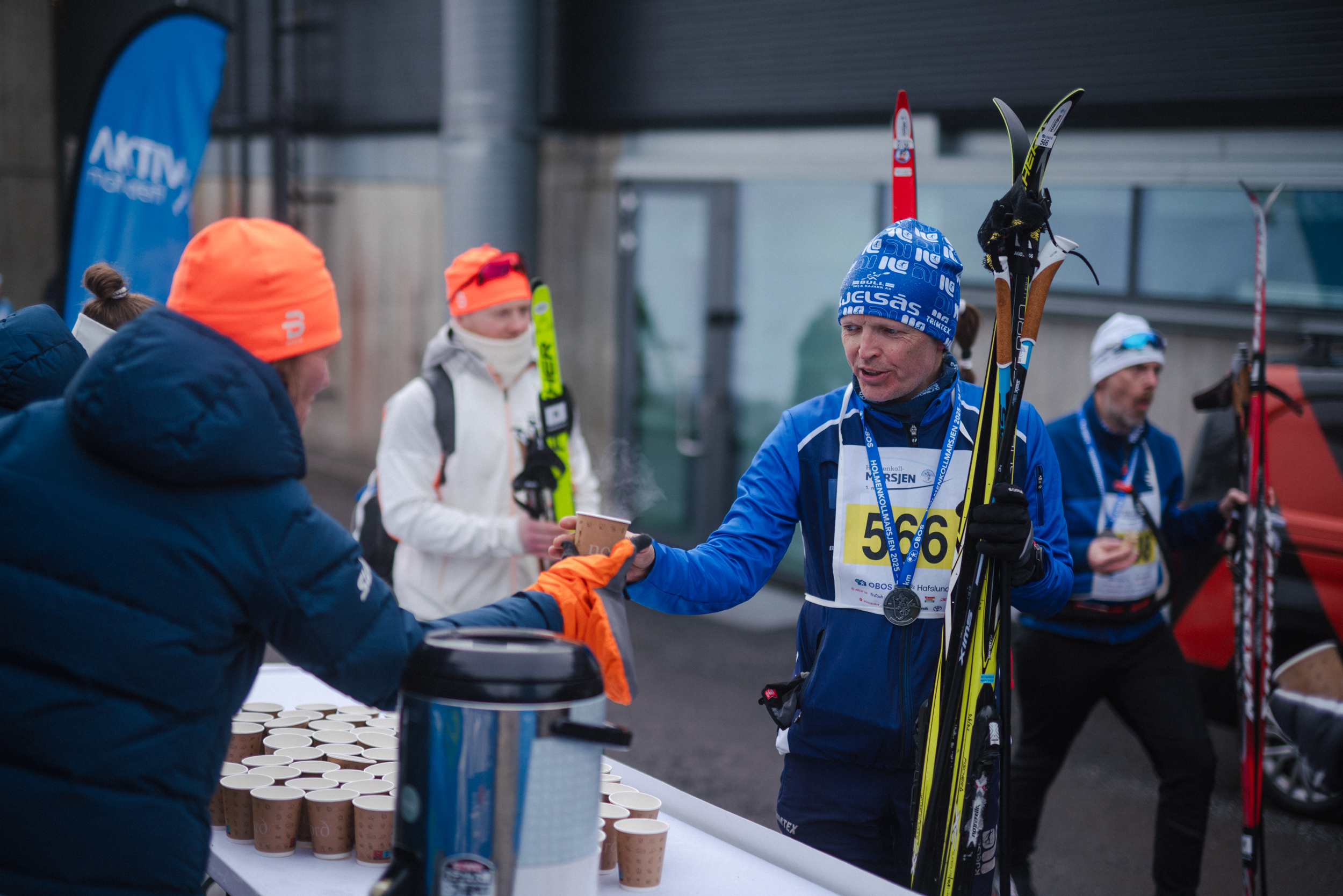 Deltakere i et skirenn mottar varme drikker ved en serveringsstasjon, med snø og skiutstyr i bakgrunnen.