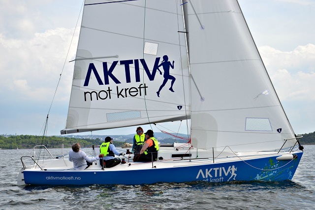 En seilbåt med logoen 'AKTIV mot kreft' seiler på sjøen, med personer ombord iført flytevester.