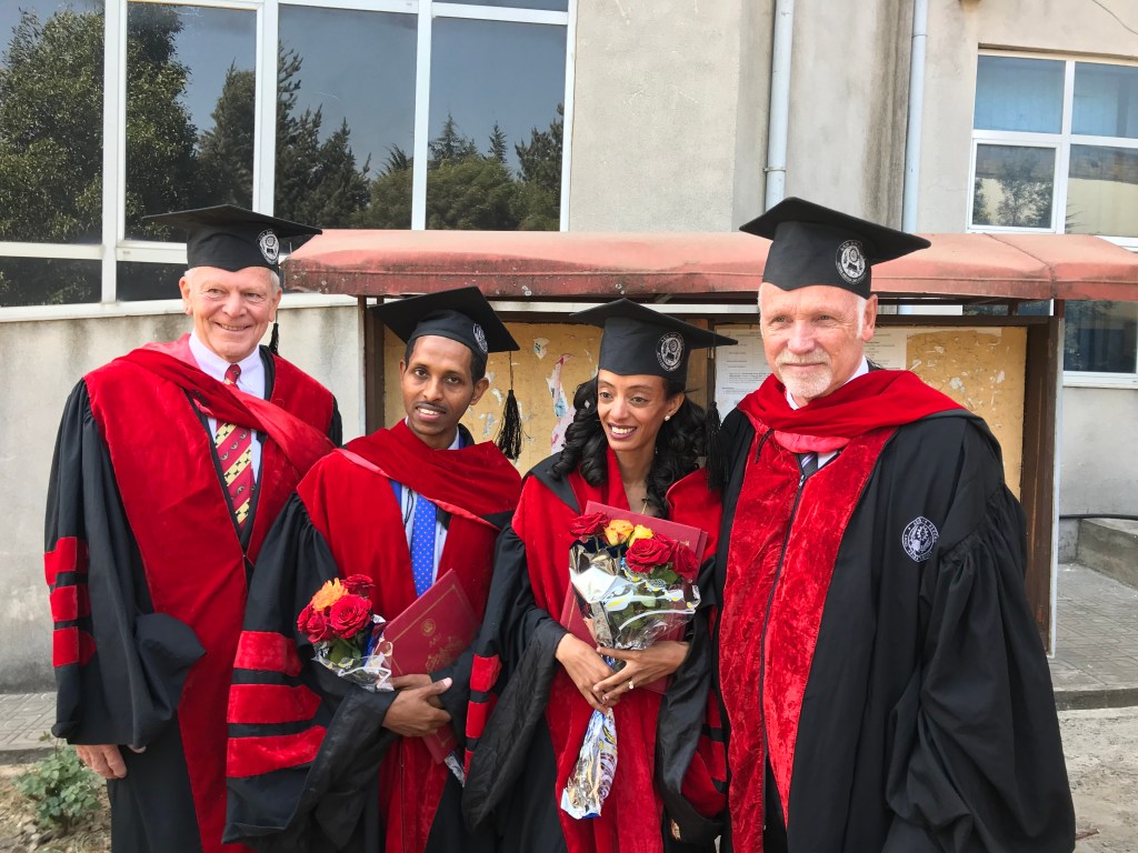 En gruppe utdannede personer iført akademiske kapper og hodeplagg, holder blomster og vitnemål, poserer foran en bygning med vinduer.