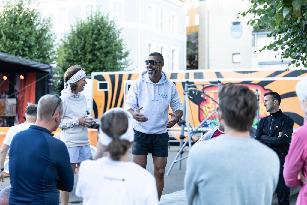 En gruppe mennesker som deltar i et utendørsarrangement, med en taler som engasjerer deltakerne. Talen skjer foran en fargerik bakgrunn og med treningsutstyr i nærheten.