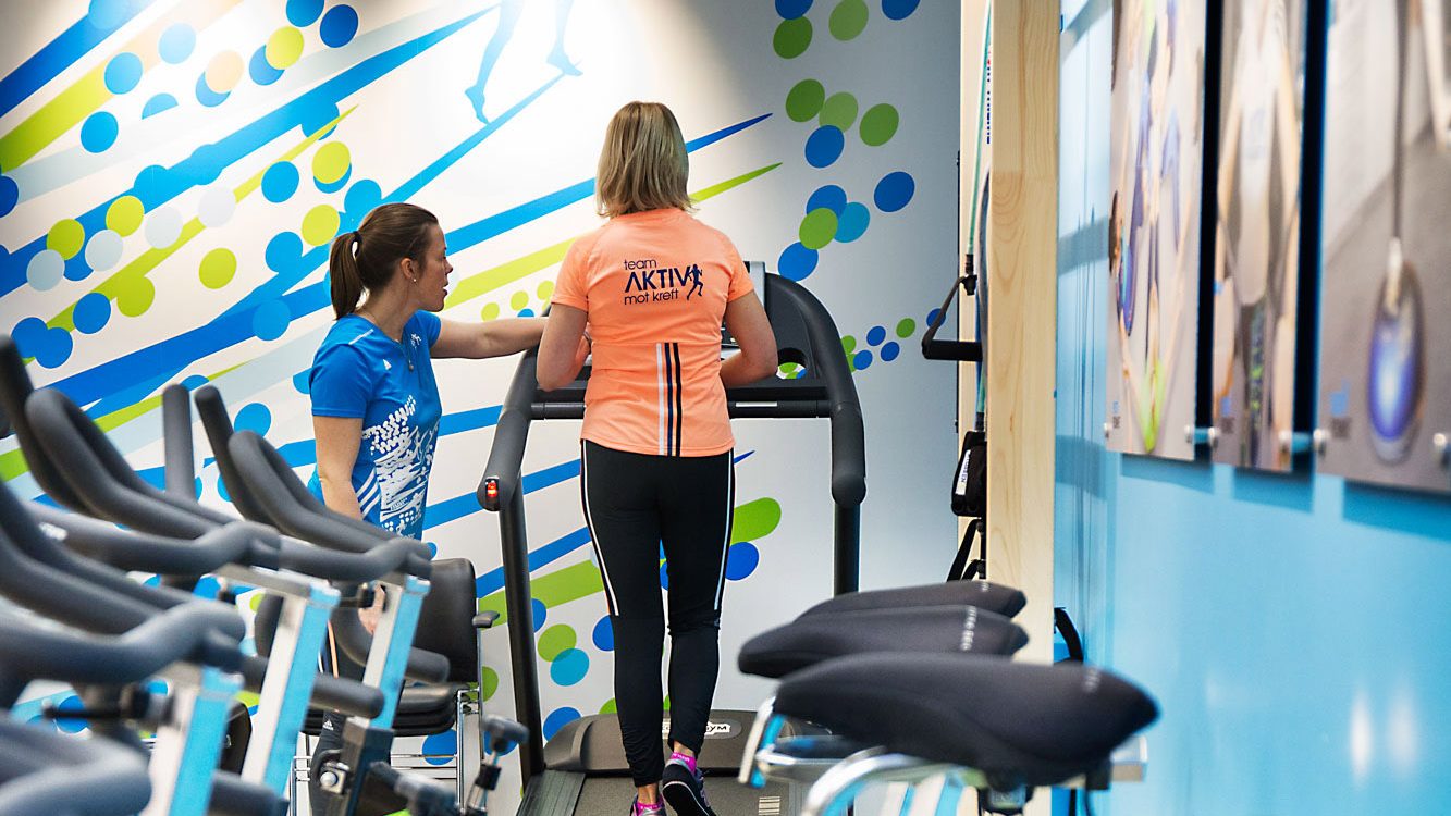 To personer trener sammen på tredemølle i et treningsstudio, med utstyr og en fargerik bakgrunn.