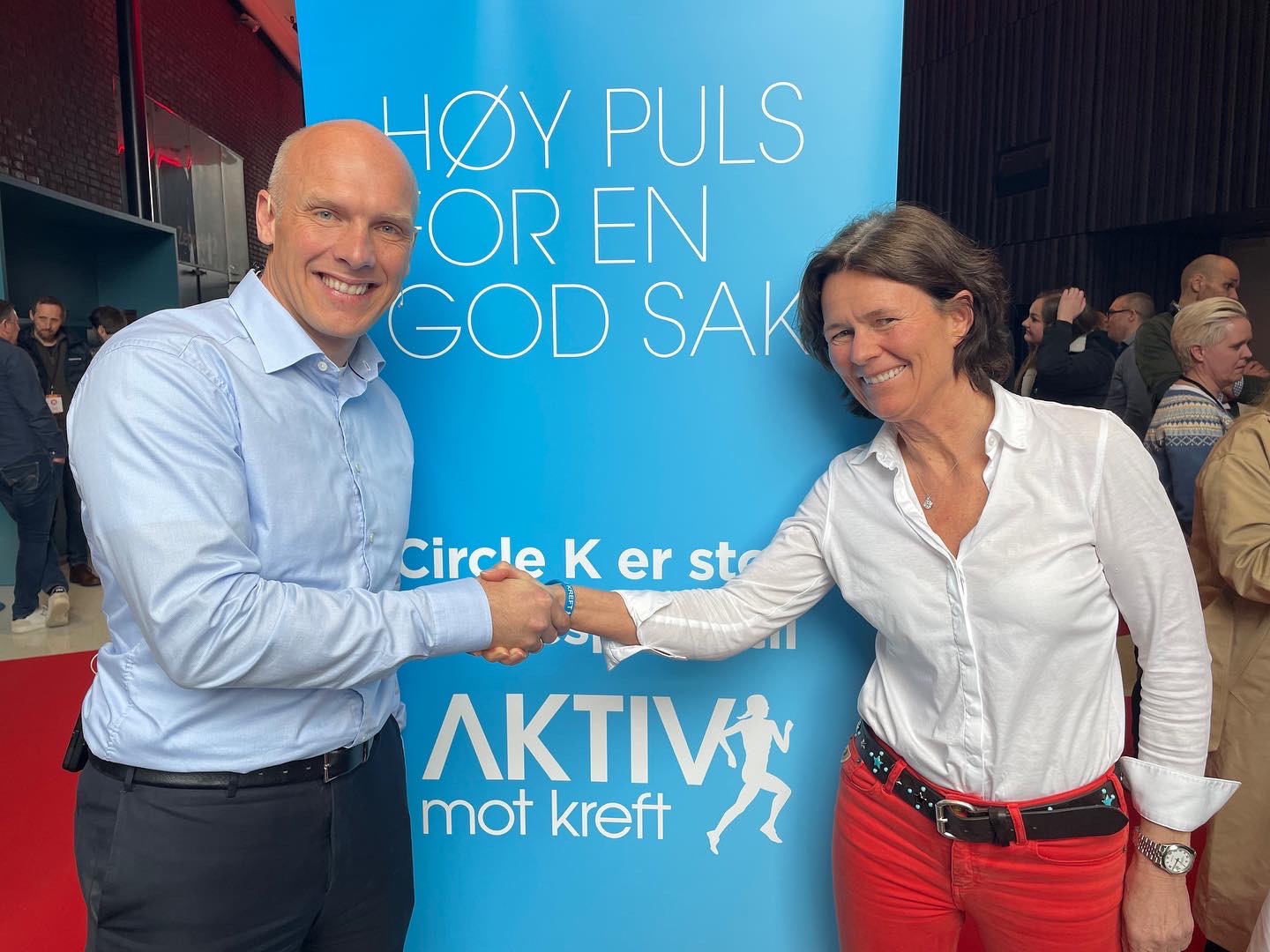 To personer som smiler og håndhilser foran en blå plakat med teksten 'HØY PULS FOR EN GOD SAK AKTIV mot kreft' og 'Circle K er stor støttespiller'.