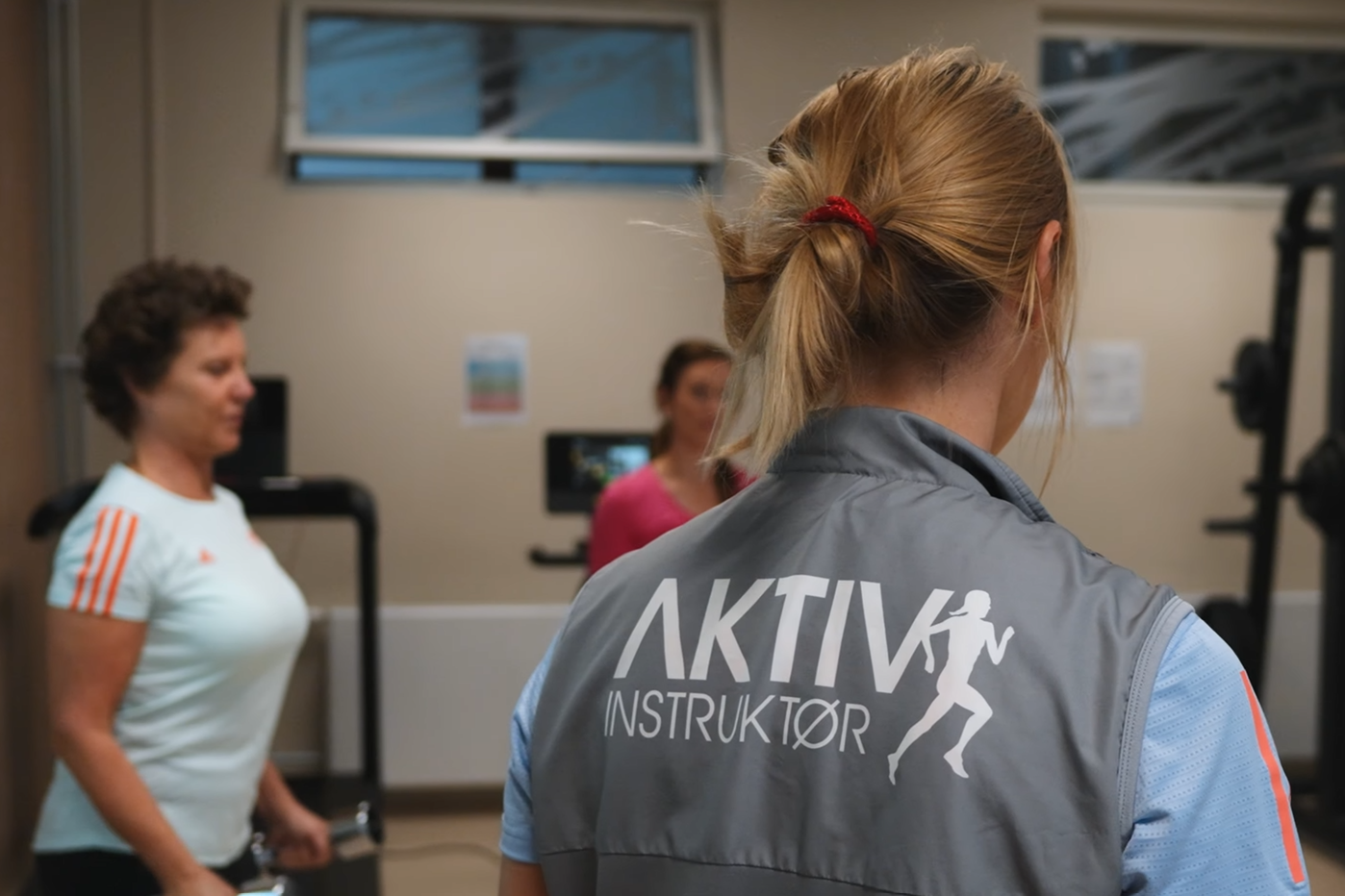 En AKTIVinstruktør leder en treningsøkt med kvinner i et treningsstudio, iført grå vest med teksten 'AKTIV INSTRUKTØR' på ryggen.