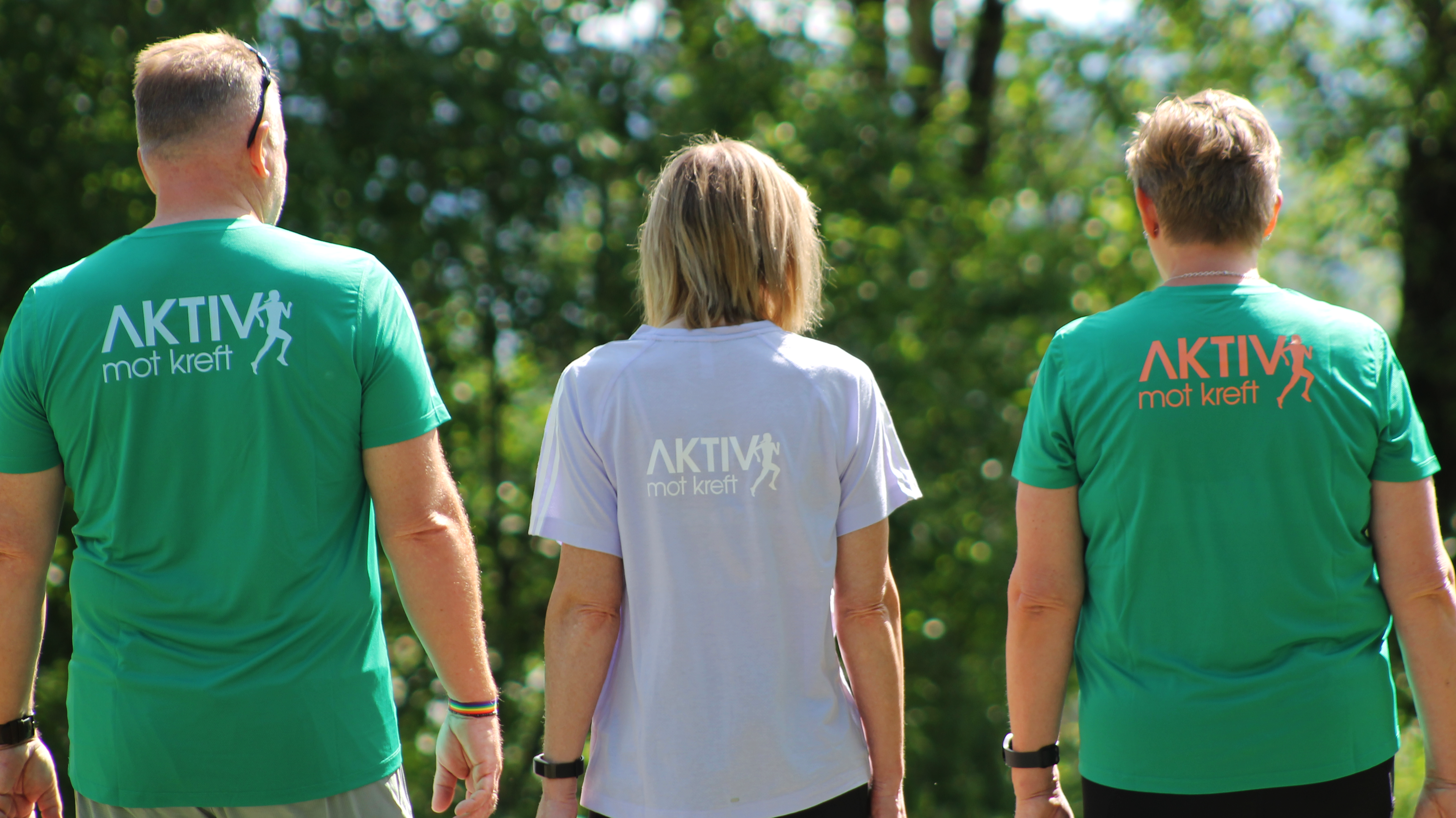 Tre personer som står med ryggen til, iført t-skjorter med teksten 'AKTIV mot kreft', i en grønn og hvit fargekombinasjon, i en grønn park med trær i bakgrunnen.